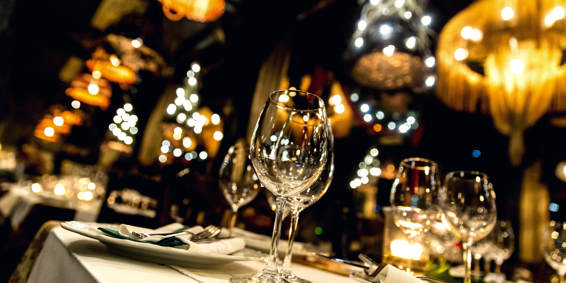 A long table laid for dinner by candlelight: glassware, folded linen and low flames running away into a dark room.