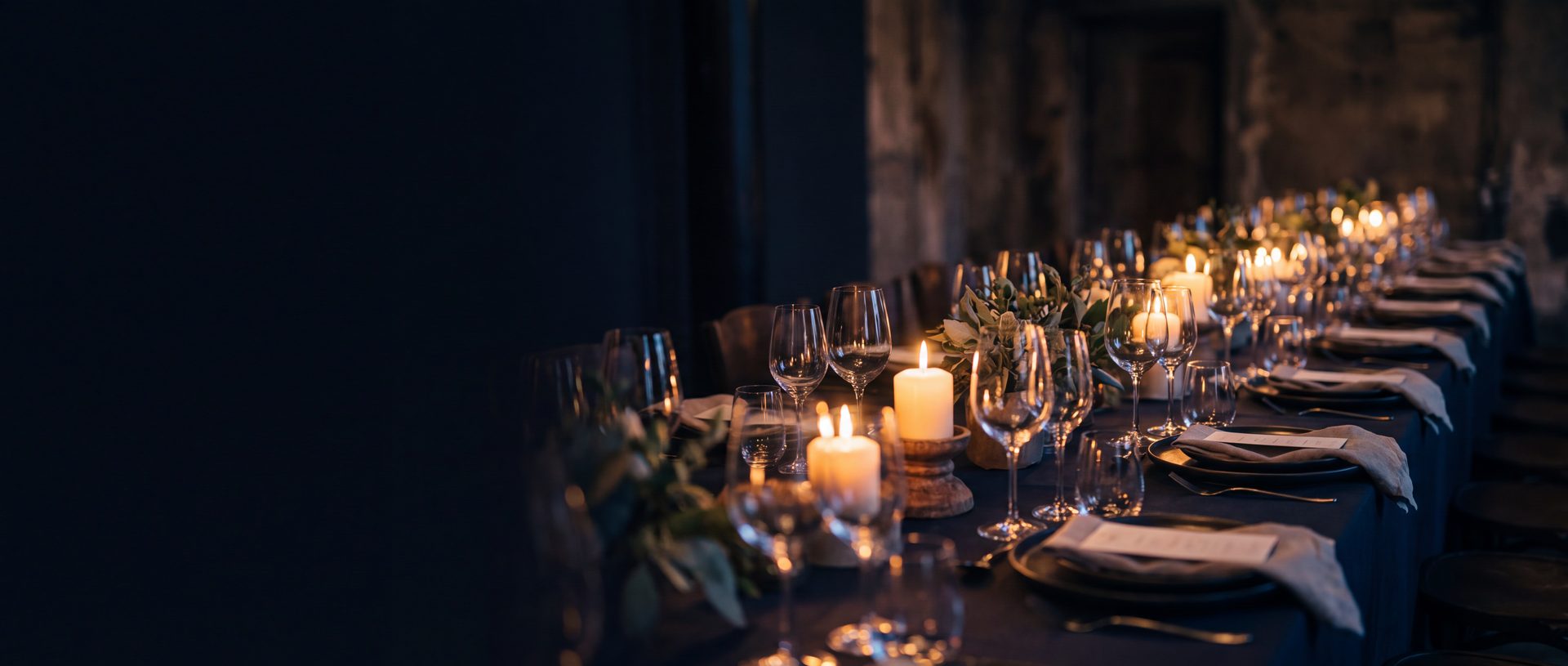 A long table laid for a private evening event: low candles down the centre, polished glassware catching the flames, dark plates and folded linen running away into a softly lit room.